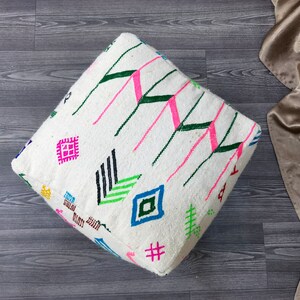 K&ouml;nnte beinhalten: Wei&szlig;er quadratischer Bodenkissen mit einem bunten geometrischen Muster. Das Muster enth&auml;lt Pink, Gr&uuml;n, Blau, Schwarz und Gelb.
