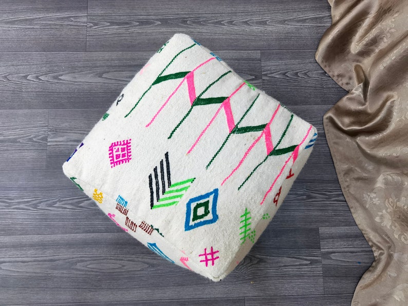 K&ouml;nnte beinhalten: Wei&szlig;er quadratischer Bodenkissen mit einem bunten geometrischen Muster. Das Muster enth&auml;lt Pink, Gr&uuml;n, Blau, Schwarz und Gelb.