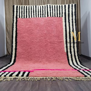 May include: A large, rectangular rug with a pink center and black and white striped borders. The rug has a textured surface and a fringe along the bottom edge. A pink horizontal stripe and a few pink rectangles are on the bottom.