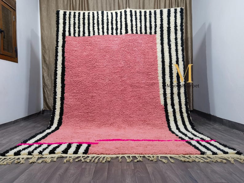 May include: A large, rectangular rug with a pink center and black and white striped borders. The rug has a textured surface and a fringe along the bottom edge. A pink horizontal stripe and a few pink rectangles are on the bottom.