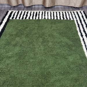 May include: A green rectangular rug with a textured surface. It features a border of alternating black and white stripes. The rug is placed on a wooden floor.