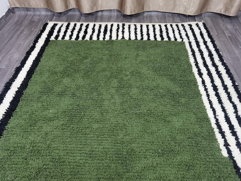 May include: A green rectangular rug with a textured surface. It features a border of alternating black and white stripes. The rug is placed on a wooden floor.