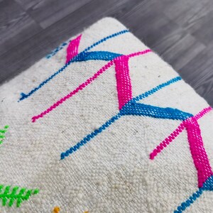 K&ouml;nnte beinhalten: Wei&szlig;es Kissen mit bunter geometrischer Stickerei. Die Stickerei zeigt rosa, blaue und gr&uuml;ne F&auml;den in einem Chevron-Muster.