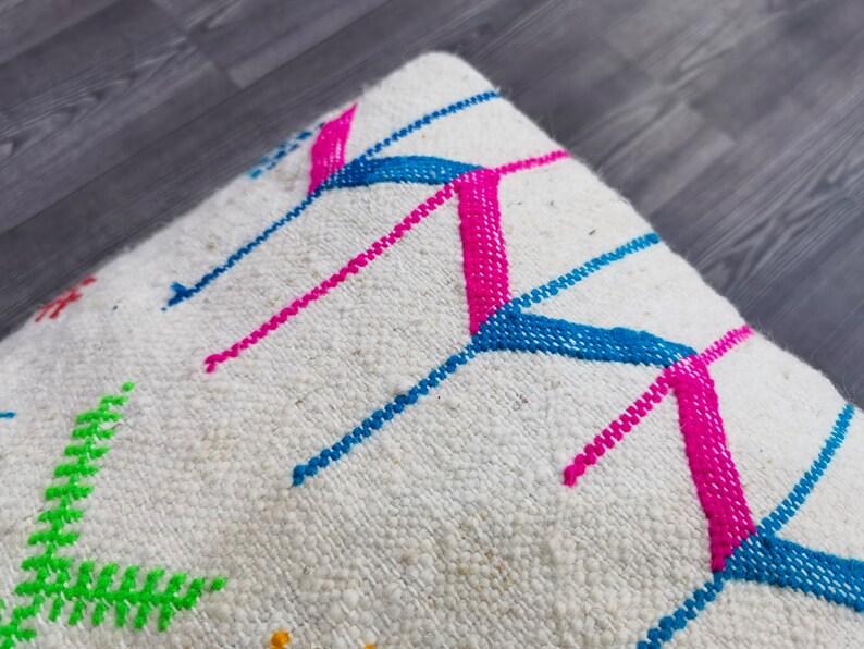 K&ouml;nnte beinhalten: Wei&szlig;es Kissen mit bunter geometrischer Stickerei. Die Stickerei zeigt rosa, blaue und gr&uuml;ne F&auml;den in einem Chevron-Muster.