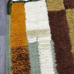 May include: A colorful, textured rug with a patchwork design. The rug features vertical stripes of mustard yellow, white, brown, and green. The top edge has a green border with white tassels. This rug is suitable for a living room or bedroom.