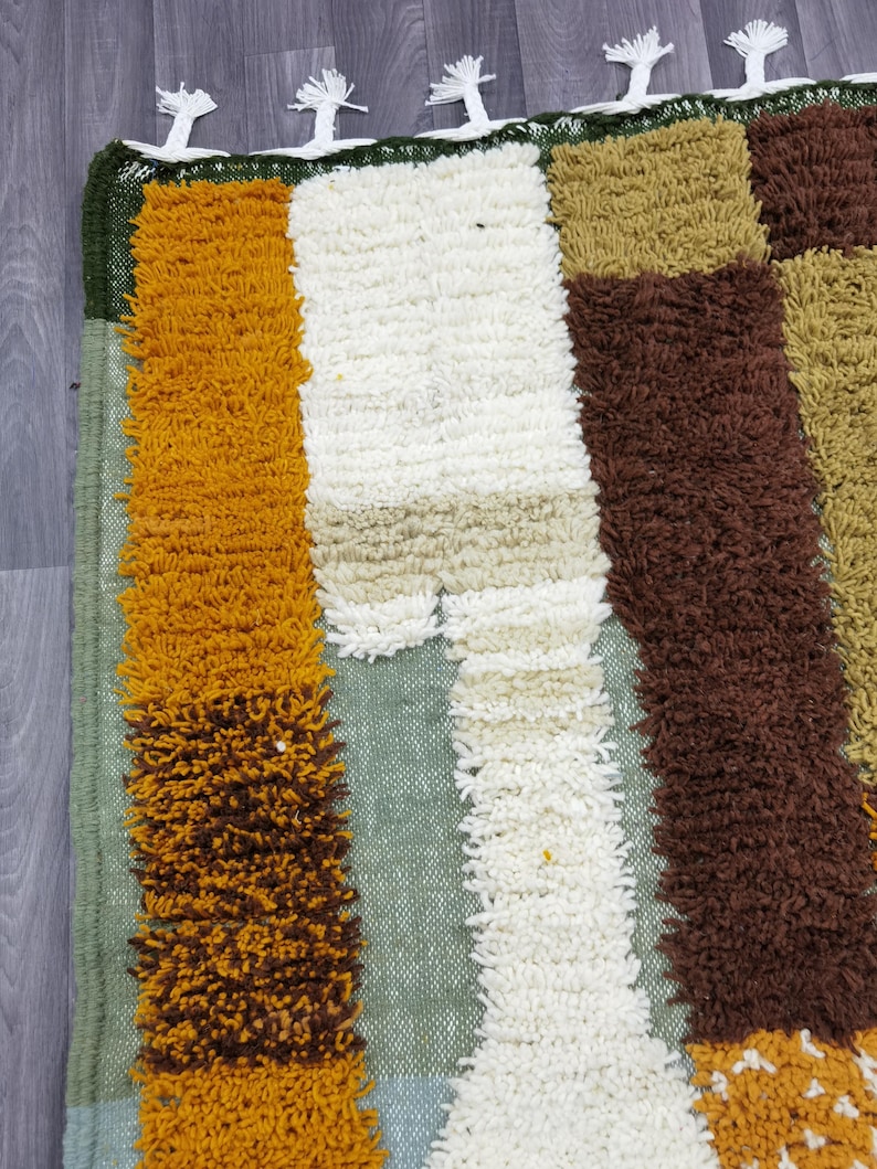 May include: A colorful, textured rug with a patchwork design. The rug features vertical stripes of mustard yellow, white, brown, and green. The top edge has a green border with white tassels. This rug is suitable for a living room or bedroom.