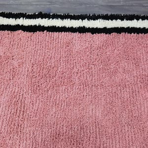 May include: A close-up of a pink rug with a textured surface. The rug has a border with a black, white, and black striped design. The rug is made of a soft, plush material.
