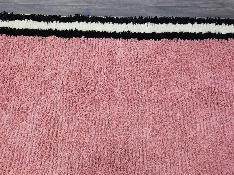 May include: A close-up of a pink rug with a textured surface. The rug has a border with a black, white, and black striped design. The rug is made of a soft, plush material.