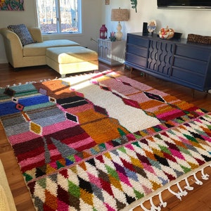 May include: A colorful Moroccan rug with a white background and geometric patterns in shades of pink, orange, blue, green, and black. The rug has white fringe along the edges.