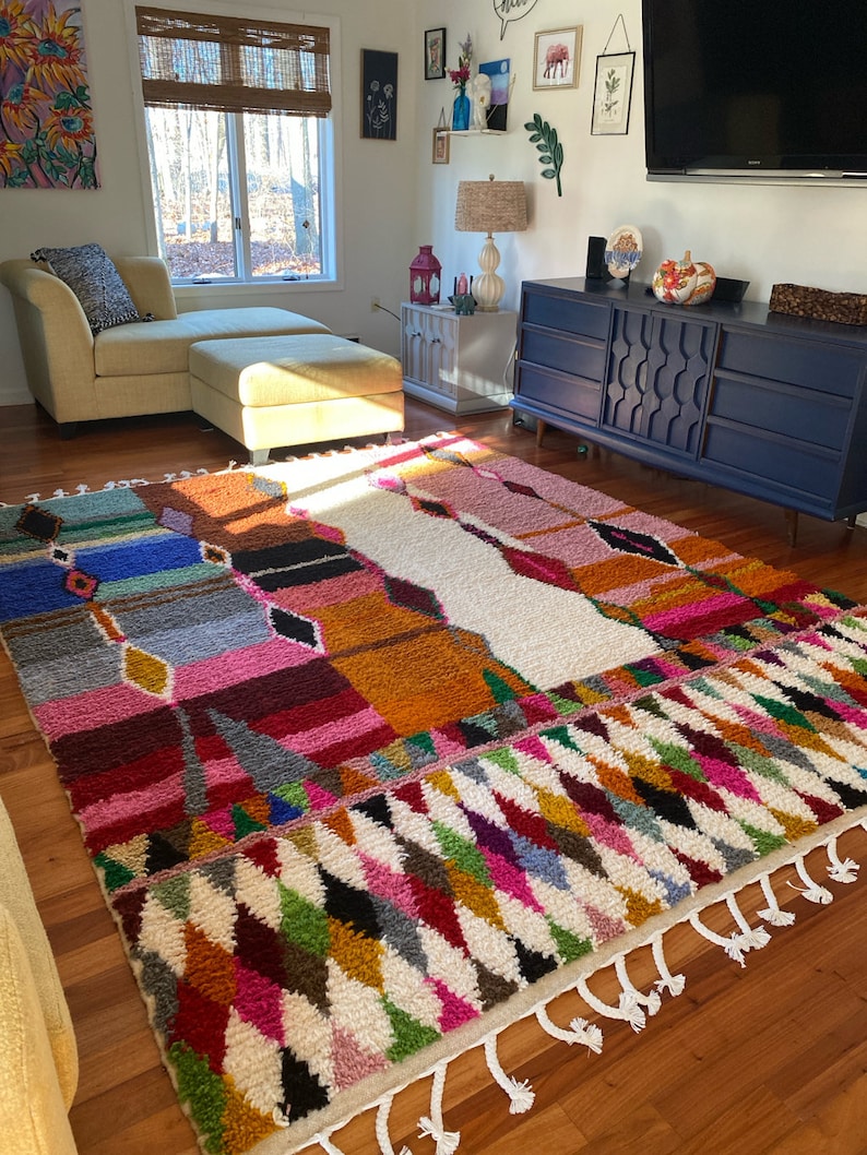 May include: A colorful Moroccan rug with a white background and geometric patterns in shades of pink, orange, blue, green, and black. The rug has white fringe along the edges.