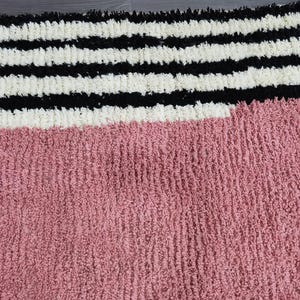 May include: A textured rug with a color block design. The top portion features horizontal stripes in black and white. The bottom section is a solid dusty rose color. The rug has a soft, plush appearance.