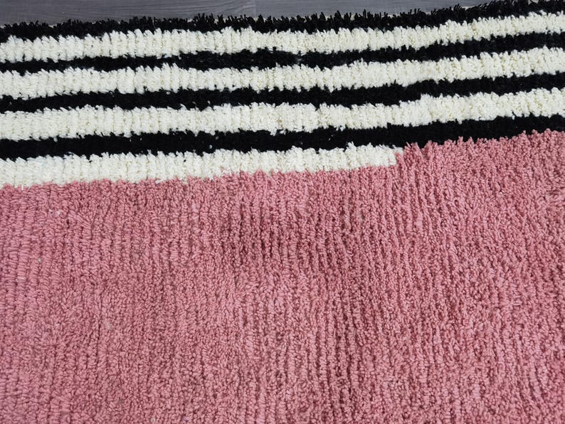 May include: A textured rug with a color block design. The top portion features horizontal stripes in black and white. The bottom section is a solid dusty rose color. The rug has a soft, plush appearance.