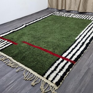 May include: Green rectangular rug with a geometric design. The rug features a green centre, a red line, and a border with black and white stripes. The rug has a fringe on the edge.