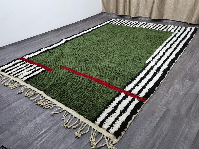 May include: Green rectangular rug with a geometric design. The rug features a green centre, a red line, and a border with black and white stripes. The rug has a fringe on the edge.