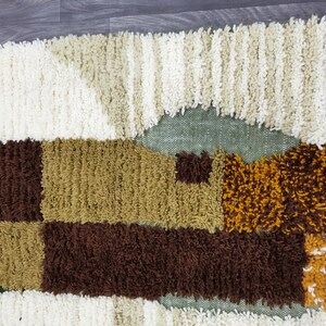 May include: A textured rug with a geometric pattern in shades of brown, tan, cream, and olive green. The rug has a shaggy, tufted texture and a fringe along one edge. The design features rectangular and triangular shapes.
