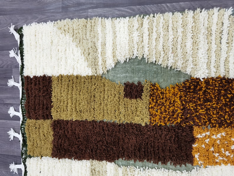 May include: A textured rug with a geometric pattern in shades of brown, tan, cream, and olive green. The rug has a shaggy, tufted texture and a fringe along one edge. The design features rectangular and triangular shapes.