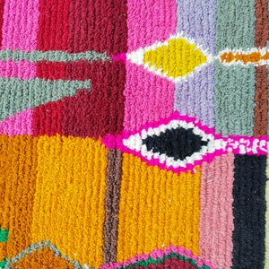 May include: A colorful rug with a geometric pattern. The rug is made of different colored yarns woven together to create a unique design. The colors include pink, red, yellow, green, blue, and black.