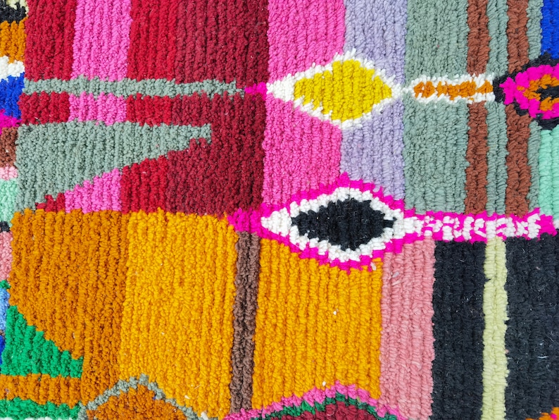 May include: A colorful rug with a geometric pattern. The rug is made of different colored yarns woven together to create a unique design. The colors include pink, red, yellow, green, blue, and black.