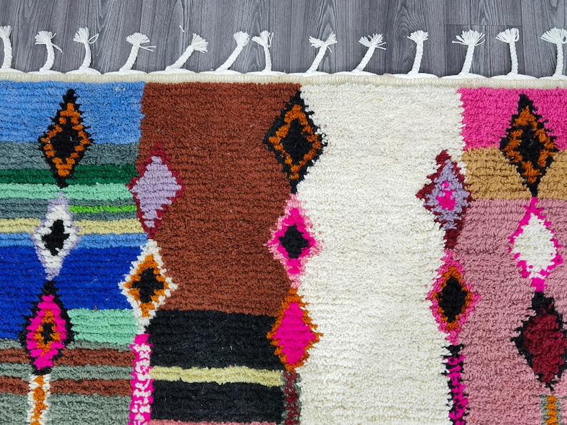 May include: A colorful Moroccan rug with a geometric pattern in shades of blue, green, pink, brown, and white. The rug has a fringe border.