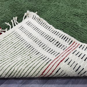 May include: A green rug with a textured surface. The rug's underside is visible, featuring a cream-coloured fabric with black and red linear patterns. The rug has a fringe along the edge.
