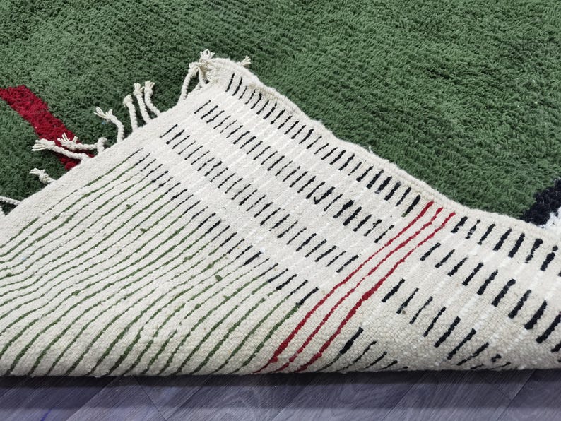 May include: A green rug with a textured surface. The rug's underside is visible, featuring a cream-coloured fabric with black and red linear patterns. The rug has a fringe along the edge.
