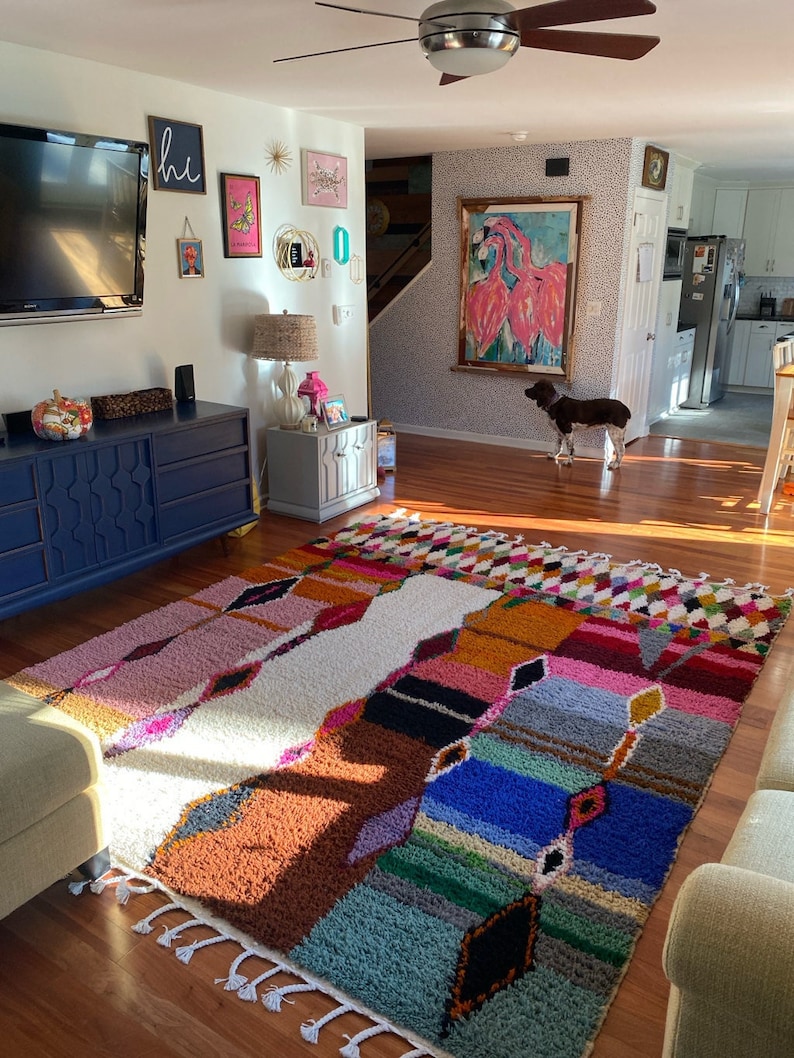 May include: A colorful, multi-patterned rug with fringe edges, laid out on a hardwood floor in a living room. The rug has a white center with pink, orange, blue, and green stripes and geometric shapes.