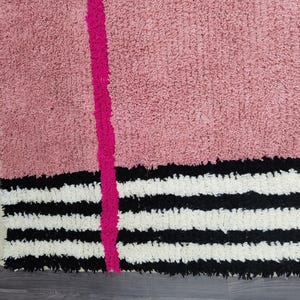 May include: A pink rug with a vertical fuchsia stripe and a black and white striped border. The rug has a textured, plush appearance and a fringe along one edge. The design is modern and geometric.