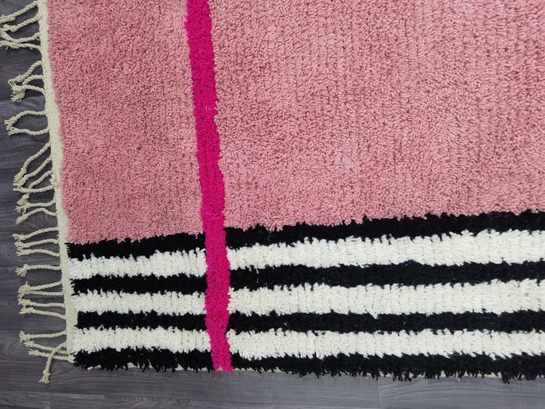 May include: A pink rug with a vertical fuchsia stripe and a black and white striped border. The rug has a textured, plush appearance and a fringe along one edge. The design is modern and geometric.