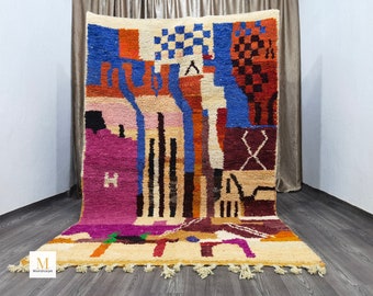 May include: A colorful, geometric Moroccan rug with a cream-colored base and a vibrant array of shapes in blue, orange, pink, and purple. The rug features a textured design and a fringe along the bottom edge. The rug is displayed on a wooden floor.