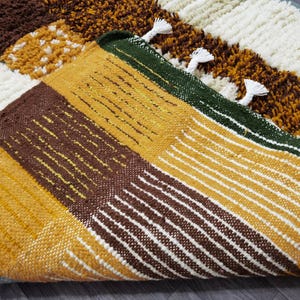 May include: Close-up of a colorful, textured rug with a geometric pattern. The rug features blocks of brown, yellow, white, and green, with white tassels along the edge. The design includes both solid and striped sections.