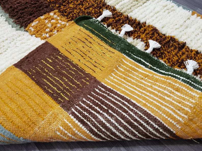 May include: Close-up of a colorful, textured rug with a geometric pattern. The rug features blocks of brown, yellow, white, and green, with white tassels along the edge. The design includes both solid and striped sections.