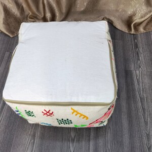 K&ouml;nnte beinhalten: Ein wei&szlig;er, quadratischer Hocker mit Rei&szlig;verschluss und bunten Stickereien. Der Hocker steht auf einem Holzboden.