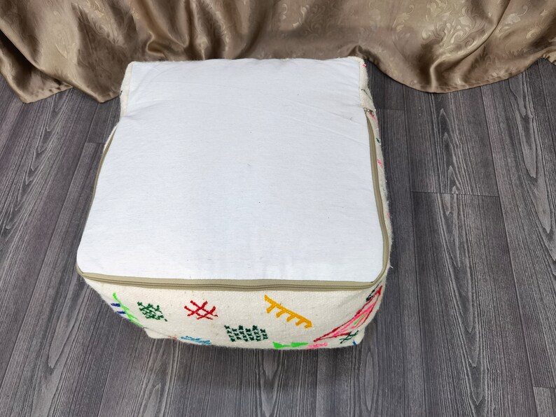 K&ouml;nnte beinhalten: Ein wei&szlig;er, quadratischer Hocker mit Rei&szlig;verschluss und bunten Stickereien. Der Hocker steht auf einem Holzboden.