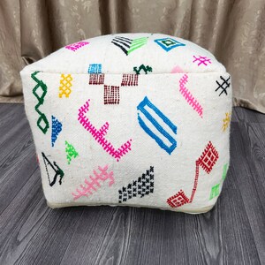 K&ouml;nnte beinhalten: Wei&szlig;er Pouf mit bunten geometrischen Mustern, die in den Stoff eingewebt sind. Der Pouf hat eine quadratische Form mit abgerundeten Kanten.