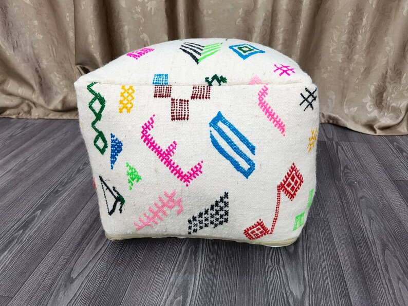 K&ouml;nnte beinhalten: Wei&szlig;er Pouf mit bunten geometrischen Mustern, die in den Stoff eingewebt sind. Der Pouf hat eine quadratische Form mit abgerundeten Kanten.