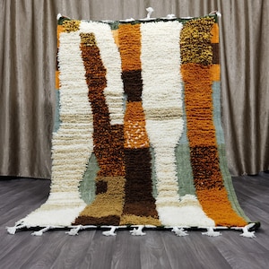 May include: A rectangular, textured rug with a patchwork design. The rug features vertical stripes in shades of white, brown, orange, and green. The rug has a fringe of white tassels along the bottom edge. The rug is approximately 5 feet by 6 feet.