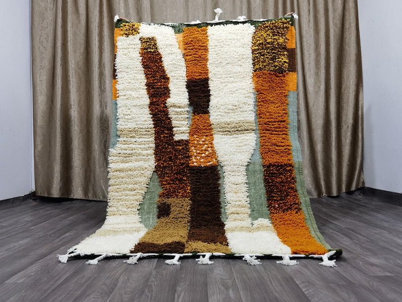 May include: A rectangular, textured rug with a patchwork design. The rug features vertical stripes in shades of white, brown, orange, and green. The rug has a fringe of white tassels along the bottom edge. The rug is approximately 5 feet by 6 feet.