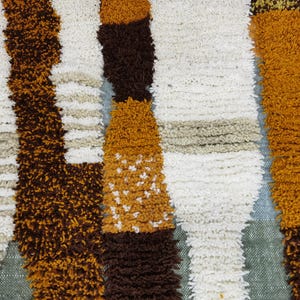 May include: Close-up of a textured rug with a repeating pattern of vertical stripes. The stripes are in shades of brown, white, tan, and gold. The rug has a soft, plush appearance, suggesting a comfortable texture.
