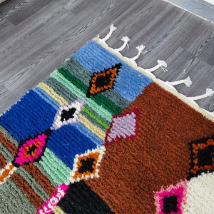 May include: A colorful Moroccan rug with a geometric pattern in shades of blue, green, brown, pink, and white. The rug has a fringe border.