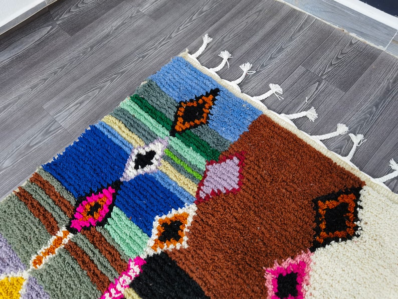 May include: A colorful Moroccan rug with a geometric pattern in shades of blue, green, brown, pink, and white. The rug has a fringe border.