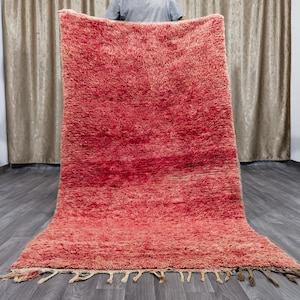 Puede incluir: Una alfombra grande, esponjosa y roja con un borde de flecos. La alfombra está doblada por la mitad y sostenida por una persona.