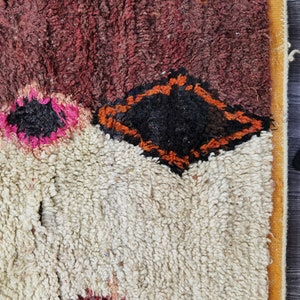 May include: A close-up of a colorful Moroccan rug with a white background and a diamond-shaped pattern in orange and black. The rug has a thick, fluffy texture.