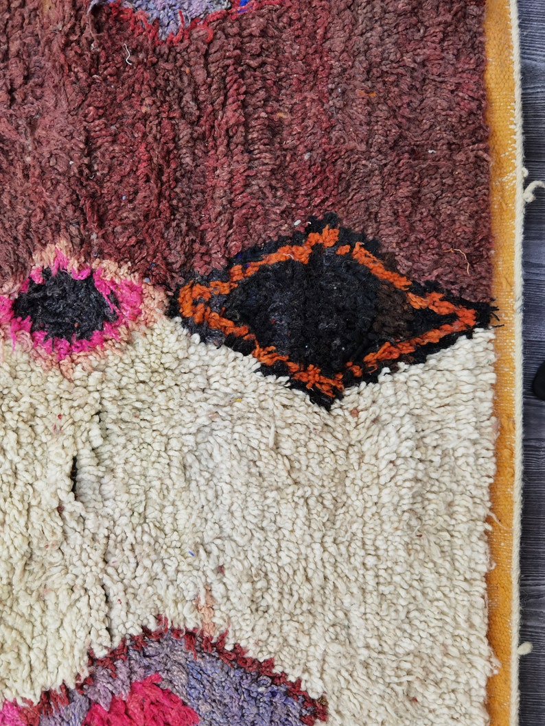 May include: A close-up of a colorful Moroccan rug with a white background and a diamond-shaped pattern in orange and black. The rug has a thick, fluffy texture.