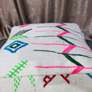 K&ouml;nnte beinhalten: Wei&szlig;er Bodenkissen mit bunten geometrischen Mustern in Rosa, Gr&uuml;n, Blau und Schwarz. Das Kissen ist aus gewebtem Stoff und hat eine quadratische Form.