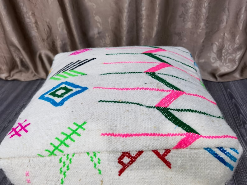 K&ouml;nnte beinhalten: Wei&szlig;er Bodenkissen mit bunten geometrischen Mustern in Rosa, Gr&uuml;n, Blau und Schwarz. Das Kissen ist aus gewebtem Stoff und hat eine quadratische Form.