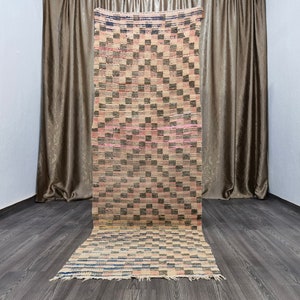 May include: A long, rectangular rug with a checkered pattern in shades of pink, black, and beige. The rug has a fringe border and is made of woven fabric.