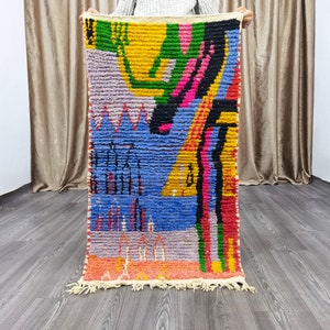 May include: A rectangular, hand-woven rug with a vibrant abstract design. The rug features a mix of colors, including yellow, blue, pink, black, and green. The design includes geometric shapes and patterns. The rug has a fringed edge and is held up by a person.