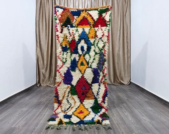 May include: A colorful Moroccan rug with a geometric pattern in shades of red, blue, yellow, green, and purple. The rug has a white background and is made of wool.
