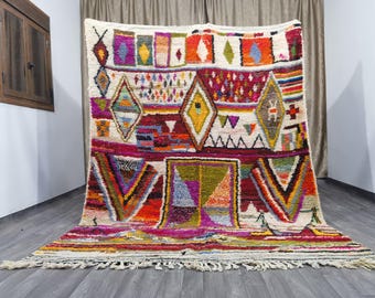 Puede incluir: Una alfombra marroquí colorida con un patrón geométrico en tonos de rojo, rosa, azul, verde y amarillo. La alfombra tiene un fondo blanco y un borde de flecos.