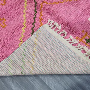 Op de afbeelding: Roze en wit geweven tapijt met een geometrisch patroon. Het tapijt heeft een franje rand en is gemaakt van zachte, pluche vezels.